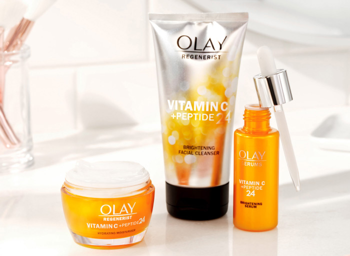'The Brighten Up Set' on a white, minimalist bathroom counter with a glass jar full of makeup brushes in the background. The set features one Olay Vitamin C + Peptide 24 Brightening Facial Cleanser, one jar of Olay Regenerist Vitamin C + Peptide 24 Moisturizer, and one Vitamin C + Peptide 24 Brightening Serum